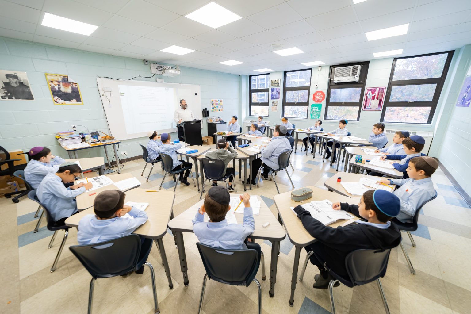 A full class of Talmidim working at modular tables in a bright classroom.